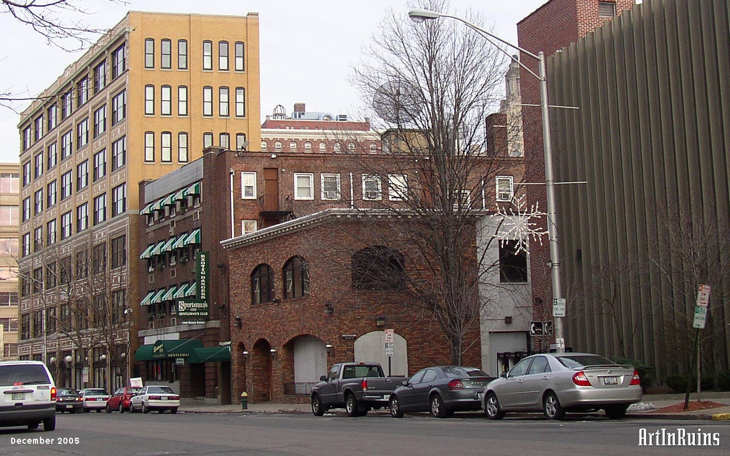 A two-story red brick facade with painted concrete block behind. Windows and door openings on the facade have arched lintels. Windows on the ground floor have been closed up with plywood.