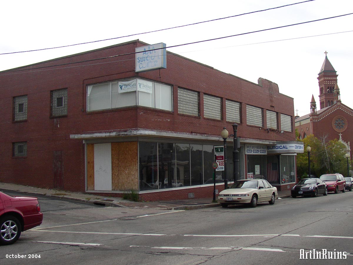 A two-story red brick building with a glass commercial storefront on the first floor. Floor plan seems basically square with an offset first floor entrance on the right side. The second story windows have been filled with glass block and a few windows in these photos are boarded up.