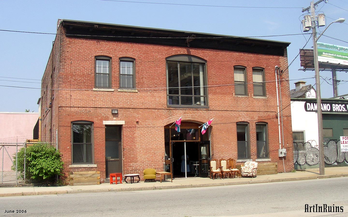 A two story red brick mill-looking building with central garage door size openings, one on each floor, flanked by sets of two arched top windows on both floors. On the first floor, an additional door entrance is on the left under the pair of windows on the second floor. Ornamentation is sparse but for a large bracketed wooden cornice along the façade’s roofline. 