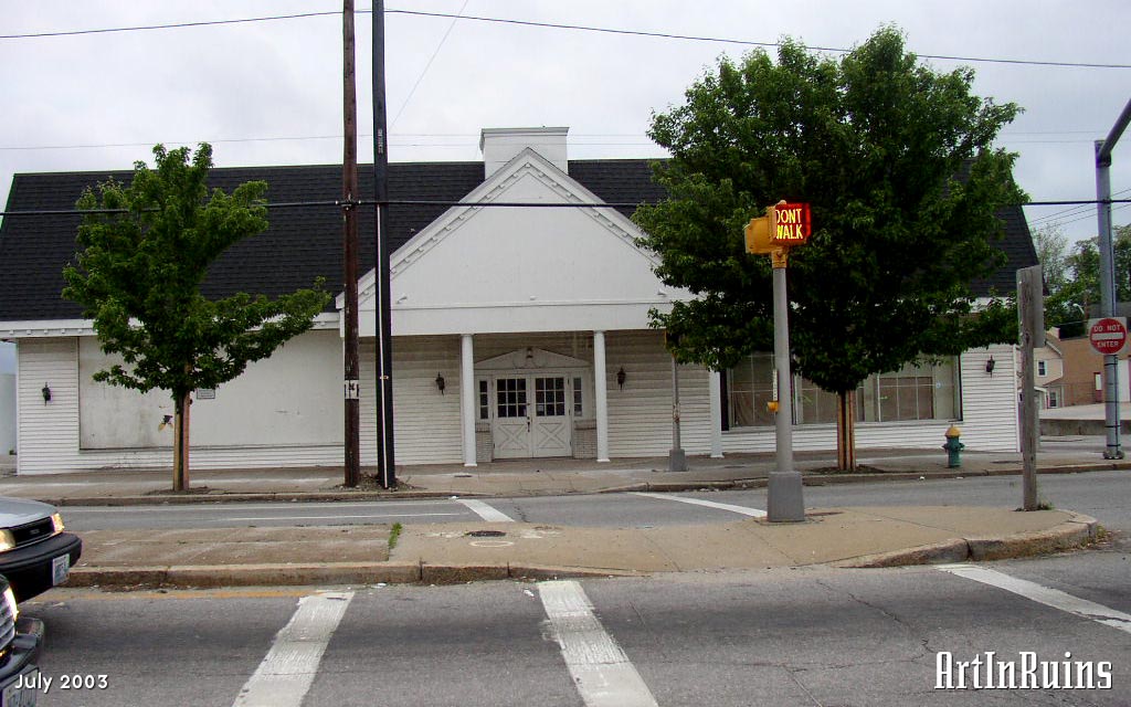 A non-descript retail building along a road of retail stores and franchises. White wooden clapboard exterior front-piece on a cinder block building. An entrance on the front with a gable roof and double commercial door underneath.