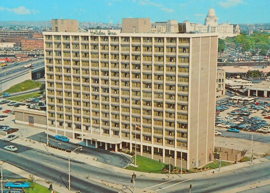 A thirteen story, simple rectangular pier and spandrel building with a high glass to coated concrete ratio. The two narrow ends of the building are blank tan brick. A central portico entrance faces Broadway while the back of the hotel is across a small street from the Providence Civic Center.