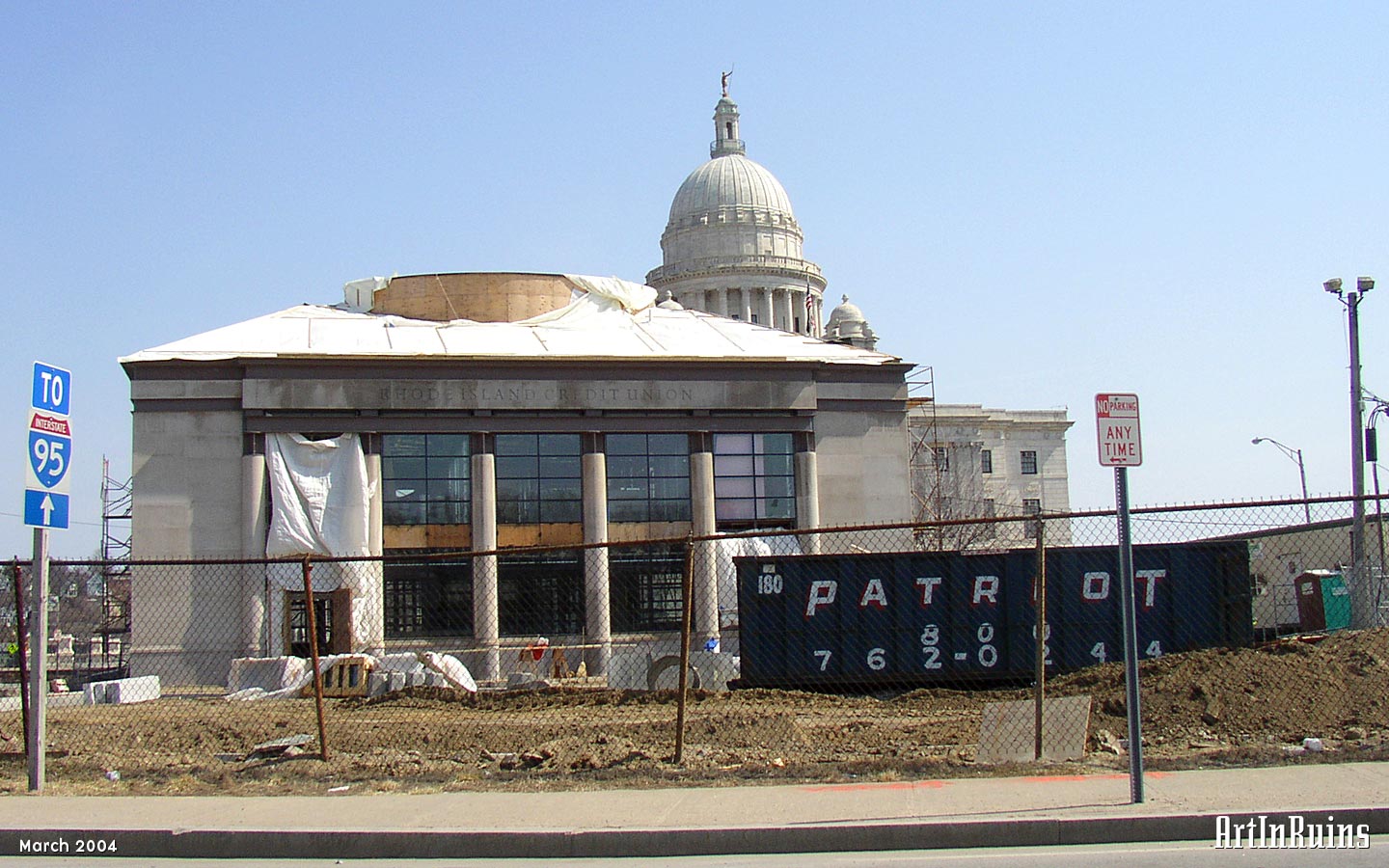 A one-story grey stone clad building with a high ceiling and central dome-like detail. Modern commercial glass panels are interspersed between classical Greek-inspired columns on two sides of the building.