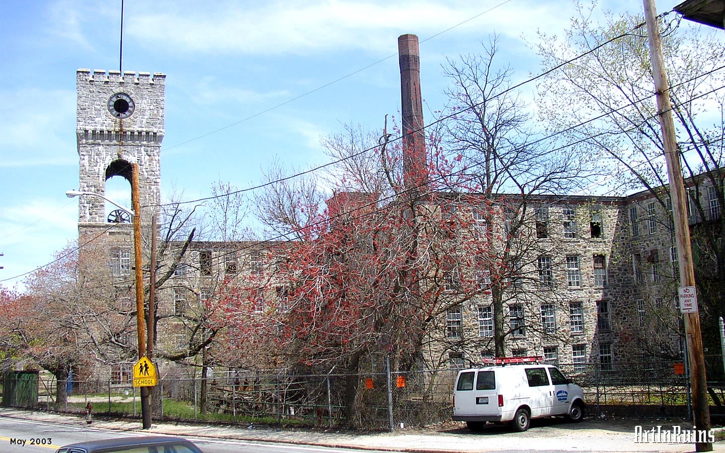 A 400 foot long stone walled mill building four stories high with impressive towers on two ends runs along Providence Street at a corner of the road where the Pawtucket River also runs. Across the river, just down from a dam, and connected by a 65 foot long truss bridge at the fourth story level, is a four-story red brick mill of relatively modern construction.