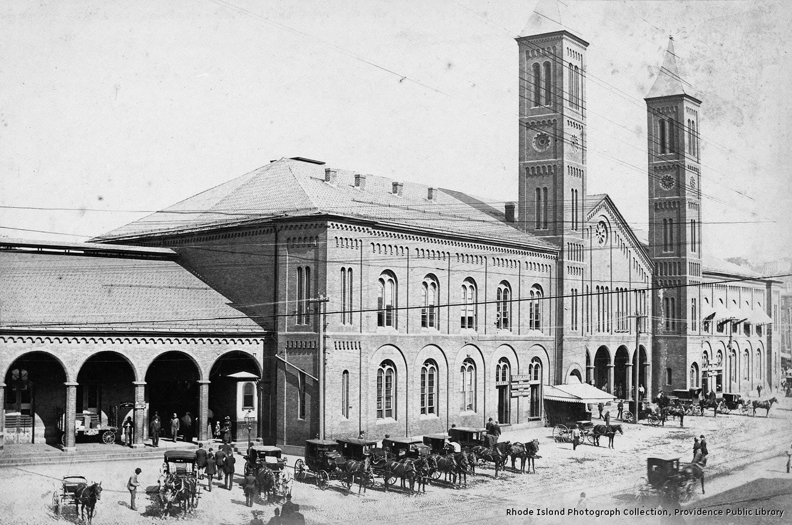A handsome red-brick railroad station with two long single story passenger terminal wings on either side of a central pavilion. Brick ornamentation was in the form of drip mouldings along the cornices and surrounding rounded arch window groupings and arched supports for covered walkway openings. A double hip roof covered the bulk of the terminal wings while the central structure resembled a church with a steep hip roof, rosette window and the peak, and two tall slender towers projecting from either side.