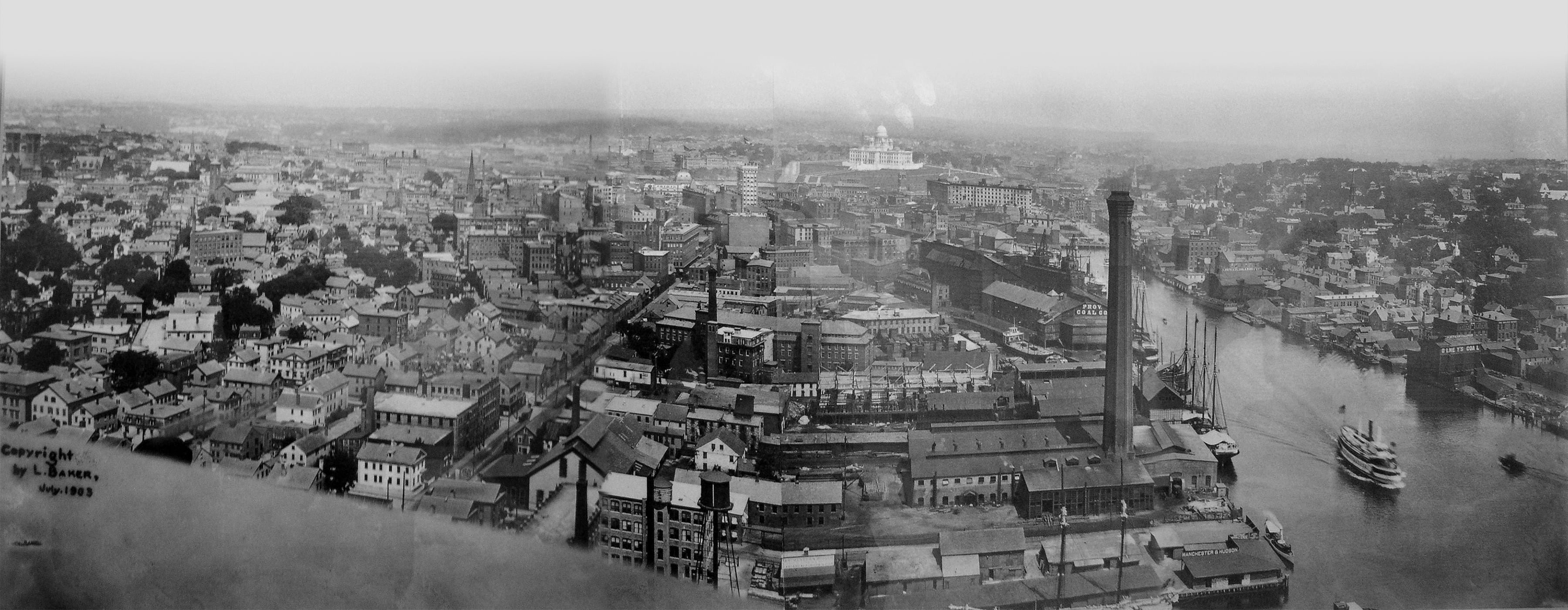 A wide panoramic black and white photo taken from a high vantage point looking north across the city of Providence at the turn of the 20th century. A dense Jewelry District is in the foreground, with the Providence River harbor on the right and the base of College Hill.