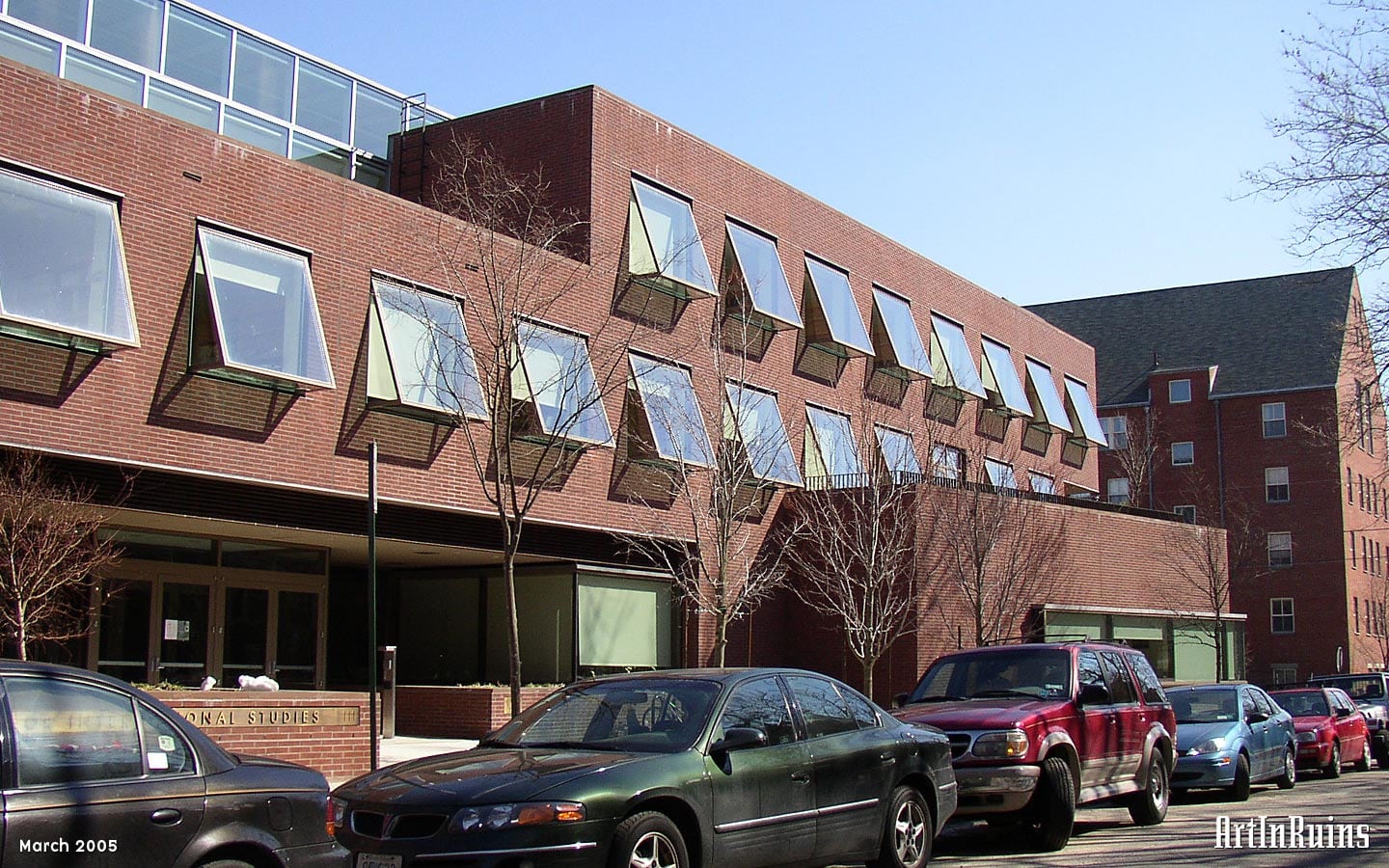 A modernist, oblong, three story brick clad building with rectangular forms and protruding awning-style windows across most of the faces. A central glass atrium and circulation area can be seen from the tall ribbon windows on either end. 